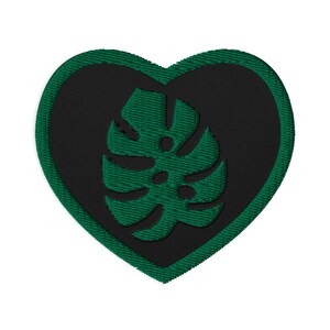 Op de afbeelding: Geborduurde patch in de vorm van een hart. De patch heeft een donkergroene rand, een zwarte hartvorm in het midden en een donkergroen monstera blad ontwerp. De patch is waarschijnlijk voor kleding of accessoires.