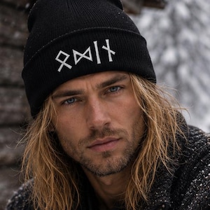 May include: A black knit beanie with white rune symbols is worn by a person with long blonde hair. The person is wearing a dark gray sweater with a patterned design. The background shows a snowy landscape and a wooden cabin.