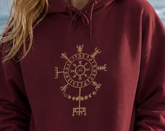 May include: A maroon hoodie with a gold-colored circular design on the front. The design features a series of symbols and lines. The hoodie has a drawstring hood and a front pocket. The person in the image is standing outdoors near the ocean.