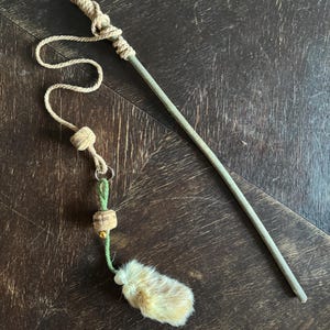 Könnte beinhalten: Ein Katzenspielzeug mit einem langen, hellgrünen Holzstab mit einem geknoteten Seilgriff. Ein Seil mit Korkakzenten und ein kleines, flauschiges, hellbraunes Fellstück hängen am Stab. Für interaktives Spiel konzipiert.