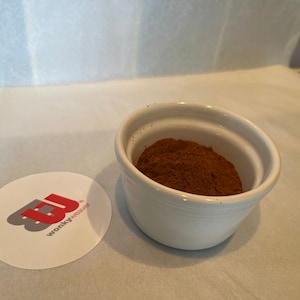 May include: A white ceramic bowl filled with a brown powder, likely cocoa powder or cinnamon, on a white surface.