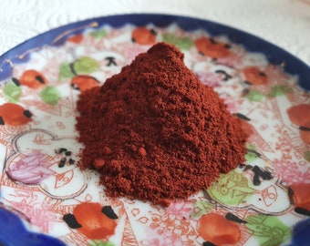 May include: A small pile of red powder on a white plate with a blue and gold rim. The plate has a floral design with red, green, and black accents.