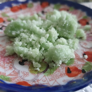May include: A close-up of a small pile of green sugar scrub on a white and red patterned plate. The scrub is made with sugar and essential oils.