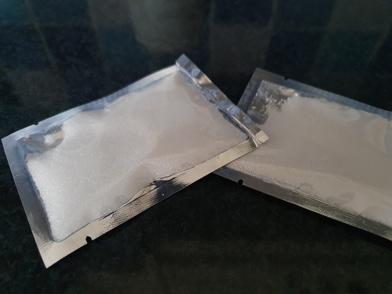 May include: Two silver foil packets filled with white powder. The packets are lying on a dark surface.