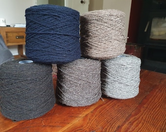 May include: Five spools of yarn in different colors: dark blue, dark gray, light gray, brown, and beige. The spools are arranged in a pyramid shape on a wooden surface.