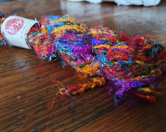Könnte beinhalten: Ein Knäuel aus buntem, mehrfarbigem, recyceltem Seidenfaden. Das Garn ist eine Mischung aus leuchtenden Farben, darunter Orange, Rot, Lila, Blau und Grün. Das Garn ist um ein weißes Etikett mit dem Text "Wool & Wander" und einem roten Kreis mit dem Buchstaben "W" darin gewickelt.