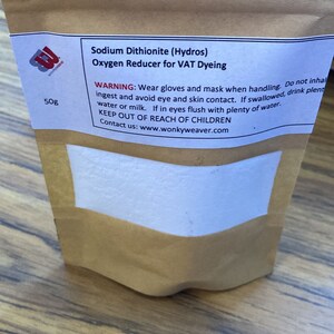 May include: A brown paper bag with a white label that reads "Sodium Dithionite (Hydros) Oxygen Reducer for VAT Dyeing". The label also includes a warning to wear gloves and a mask when handling the product, and to keep it out of reach of children. The bag contains a white powder and is labeled with a weight of 50g.
