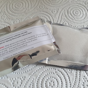 May include: A white, resealable pouch containing a white powder labeled "Aluminium potassium sulphate (ALUM) Mordant for dyeing fibres with natural dyes". The label also includes a warning about handling the powder and contact information for the company.