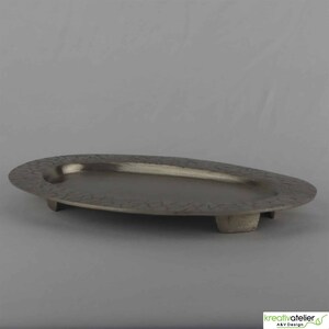 Peut inclure: Un plateau de service ovale gris foncé avec un bord texturé et trois petits pieds. Le plateau a une surface lisse et métallique et un design minimaliste, adapté pour servir des apéritifs ou exposer de petits objets.