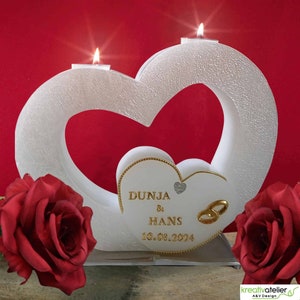 Puede incluir: Un portavelas blanco en forma de corazón con dos velas encendidas. El soporte tiene una superficie texturizada y un corazón más pequeño con los nombres "DUNJA & HANS" y la fecha "10.08.2024". Dos rosas rojas a cada lado.