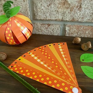 Pode incluir: Um projeto de artesanato com uma ab&oacute;bora de papel com listras laranja e amarelas e bolinhas. A ab&oacute;bora &eacute; feita de tiras de papel e tem um caule verde. H&aacute; tamb&eacute;m um leque de tiras de papel com listras laranja, amarelas e brancas e bolinhas. O leque &eacute; feito de tiras de papel e tem um bot&atilde;o branco na ponta.