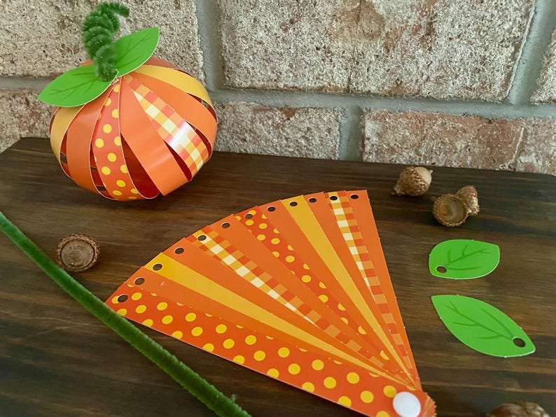 Pode incluir: Um projeto de artesanato com uma ab&oacute;bora de papel com listras laranja e amarelas e bolinhas. A ab&oacute;bora &eacute; feita de tiras de papel e tem um caule verde. H&aacute; tamb&eacute;m um leque de tiras de papel com listras laranja, amarelas e brancas e bolinhas. O leque &eacute; feito de tiras de papel e tem um bot&atilde;o branco na ponta.