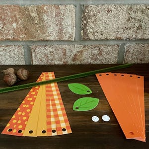 Pode incluir: Um kit de artesanato para fazer uma decora&ccedil;&atilde;o de ab&oacute;bora. O kit inclui tiras de papel com padr&otilde;es laranja e amarelo, folhas de feltro verde, olhos saltitantes brancos, um limpador de cachimbo verde e um pau marrom.