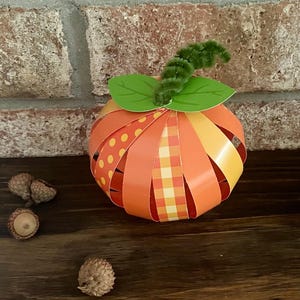 Pode incluir: Uma ab&oacute;bora de papel com um caule verde e listras de padr&otilde;es laranja, amarelo e branco. A ab&oacute;bora est&aacute; sobre uma superf&iacute;cie de madeira com bolotas.