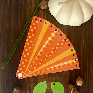 Pode incluir: Um kit de artesanato de outono com um leque de papel estampado laranja e amarelo, duas folhas verdes e bolotas.
