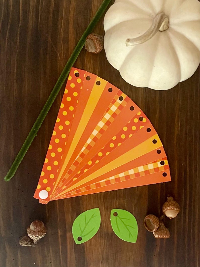 Pode incluir: Um kit de artesanato de outono com um leque de papel estampado laranja e amarelo, duas folhas verdes e bolotas.