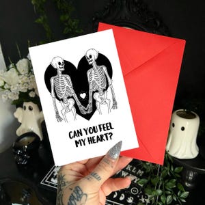 Puede incluir: Una tarjeta blanca con un diseño de corazón negro con dos esqueletos tomados de la mano, con el texto "CAN YOU FEEL MY HEART?" Un sobre rojo está detrás de la tarjeta. La tarjeta se sostiene en una mano con una uña brillante.