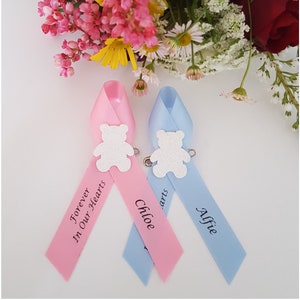 May include: Two satin ribbons, one pink and one blue, each with a white glitter teddy bear pin. The pink ribbon reads "Forever In Our Hearts" and "Chloe", while the blue ribbon reads "Alfie". A bouquet of flowers is in the background.