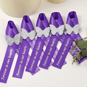 May include: Five purple satin awareness ribbons arranged in a row, each featuring a silver glitter butterfly accent and the white printed text "Forever in our hearts" along with additional dates printed on the lower portion of the ribbon tails.