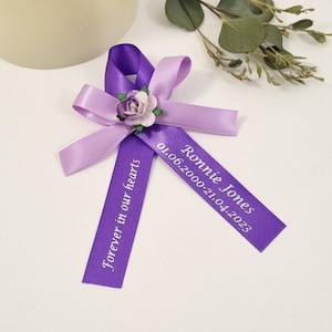 May include: Purple and lavender ribbon tied in a bow with a small flower embellishment. The ribbon has the text "Ronnie Jones 01.06.2000-21.04.2023" and "Forever in our hearts" printed on it.