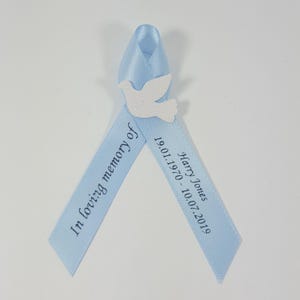 May include: A light blue satin ribbon folded into a loop, featuring a white glittery dove cutout at the centre. The left tail is printed with the text In loving memory of, and the right tail displays the name Harry Jones and dates 19.01.1970 - 10.07.2019.