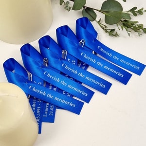 May include: A set of five royal blue satin ribbon pins, each featuring a silver safety pin and the printed text "Cherish the memories" along with individual names and dates, arranged in a diagonal row on a plain white surface.