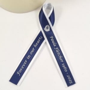 May include: A navy blue ribbon with white edging, featuring the text "Forever in our hearts" and "Paula Fletcher 1960 - 2025". The ribbon also has a small emblem. This ribbon is likely used for remembrance or memorial events.