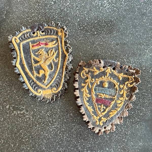 May include: Two shield-shaped leather patches with scalloped edges. The left patch features a gold-painted lion holding a flag on a dark background. The right patch displays a central crest with horizontal bands in blue, white, and red.