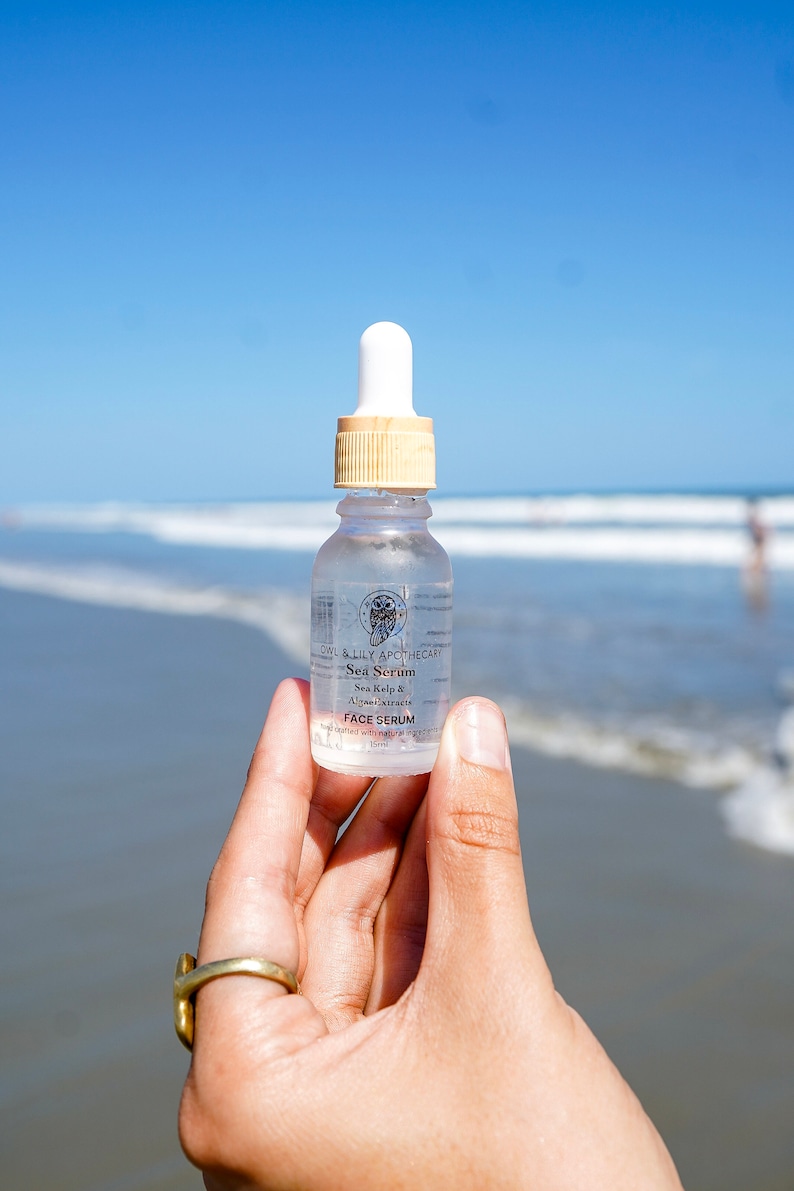 May include: A clear glass bottle of Owl & Lily Apothecary Sea Serum face serum with a dropper top. The bottle is labeled with the product name, ingredients, and a small owl logo. The bottle is being held in a hand against a blurred background of a beach.