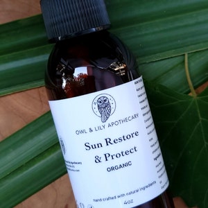 Könnte beinhalten: Eine braune Glas-Sprühflasche mit schwarzem Deckel, beschriftet mit "Sun Restore & Protect ORGANIC" von Owl & Lily Apothecary. Die Flasche enthält 118 ml Produkt und steht vor grünem Laub und Holz.