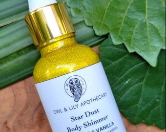 May include: A small glass bottle with a gold lid and a yellow shimmery liquid inside. The bottle is labeled "OWL & LILY APOTHECARY Star Dust Body Shimmer VITAMIN E & VANILLA hand crafted with natural ingredients 30ml".