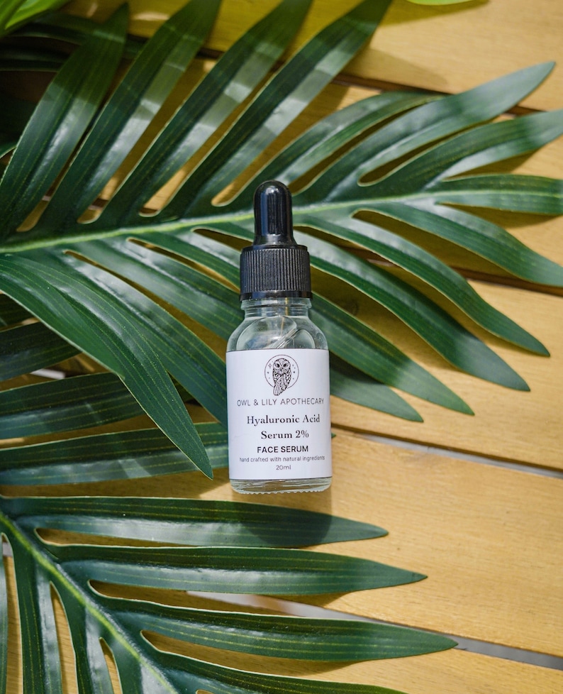 May include: A clear glass bottle with a black dropper top and a white label. The label reads "OWL & LILY APOTHECARY Hyaluronic Acid Serum 2% FACE SERUM hand crafted with natural ingredients 20ml".