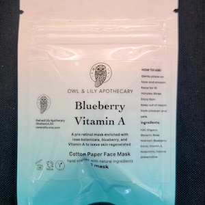 Puede incluir: Una bolsa blanca con un diseño azul y verde. La bolsa contiene una mascarilla facial de un solo uso. El texto en la bolsa dice "Blueberry Vitamin A Cotton Paper Face Mask hand crafted with natural ingredients 1 mask".