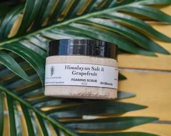 Puede incluir: Un tarro blanco con tapa negra que contiene exfoliante espumoso de sal del Himalaya y pomelo. El tarro está etiquetado con el texto "Himalayan Salt & Grapefruit Foaming Scrub 4 oz. 84 Minerals". El tarro está sobre una superficie de madera con una planta verde frondosa en el fondo.