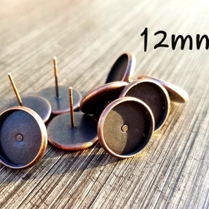 May include: A collection of antique bronze-colored earring posts with flat, round settings. The posts have a small pin for insertion and are approximately 12mm in diameter. The image shows several pieces scattered on a wooden surface.