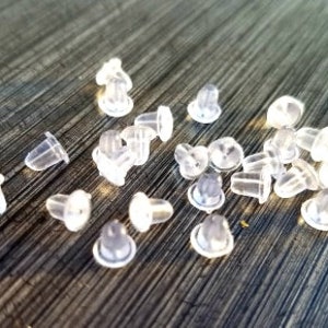 May include: A collection of clear, translucent earring backs scattered on a dark, textured surface. The earring backs are small, with a variety of shapes, including mushroom and bullet styles. The image is well-lit, highlighting the details of the earring backs.