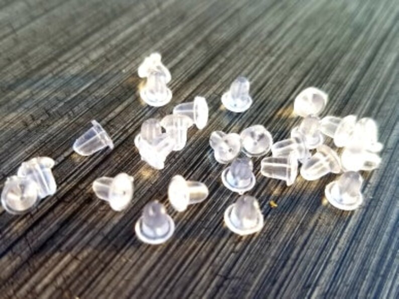 May include: A collection of clear, translucent earring backs scattered on a dark, textured surface. The earring backs are small, with a variety of shapes, including mushroom and bullet styles. The image is well-lit, highlighting the details of the earring backs.