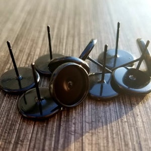 May include: A collection of black earring posts with flat, round bases and thin, straight posts. The posts are scattered on a wooden surface, with some in focus and others slightly blurred. The overall aesthetic is simple and modern, suitable for jewelry making.