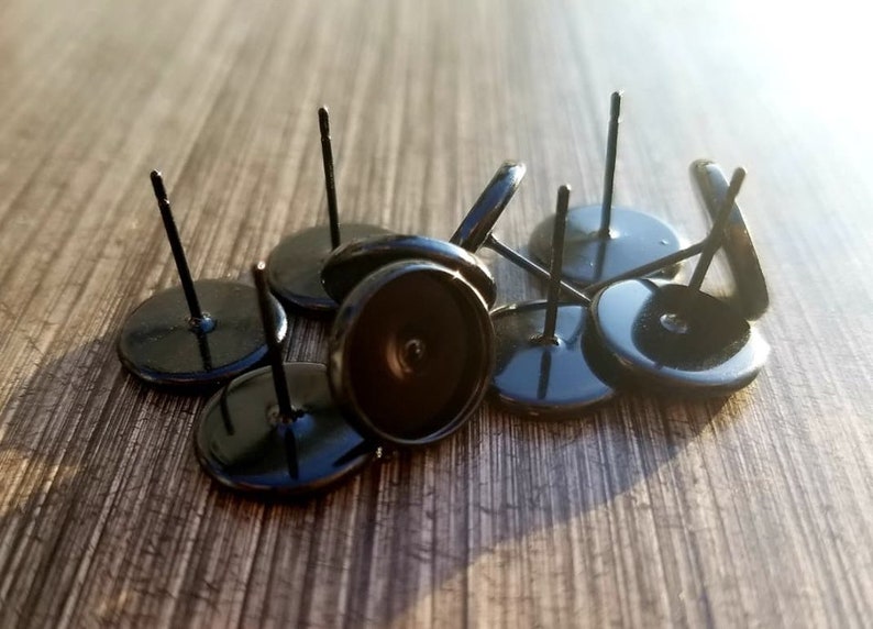 May include: A collection of black earring posts with flat, round bases and thin, straight posts. The posts are scattered on a wooden surface, with some in focus and others slightly blurred. The overall aesthetic is simple and modern, suitable for jewelry making.