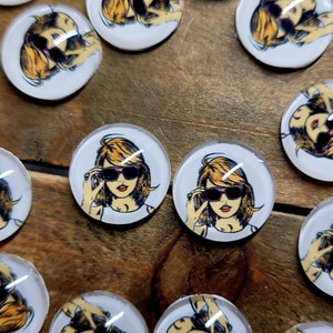 May include: A set of round, white, glass cabochons with a cartoon image of a woman wearing sunglasses. The woman has blonde hair and is wearing a white shirt.