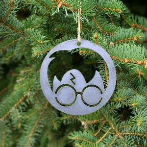 May include: A metal Christmas ornament with a circular design. The ornament features a silhouette of glasses, a lightning bolt, and a shape resembling a bird or egg. It hangs from a natural twine string against a backdrop of green pine needles.