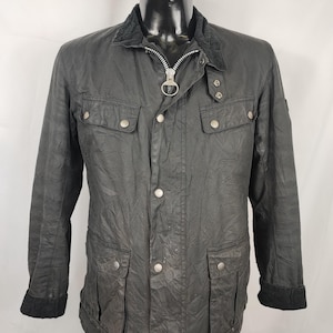 Può includere: Giacca in cotone cerato grigio scuro con colletto in velluto a coste nero, cerniera argentata e tasche frontali multiple. La giacca ha bottoni argentati e polsini neri. La giacca è esposta su un manichino nero.