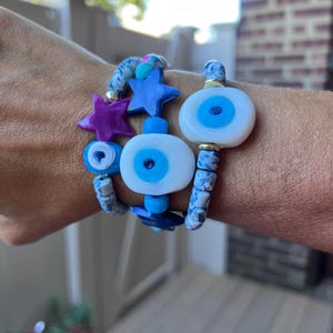 Op de afbeelding: Drie armbanden met blauwe en witte kralen. Eén heeft een grote wit en blauw oogvormige kraal, een andere heeft blauwe stervormige kralen en een paarse ster. De armbanden zijn op een pols gestapeld.