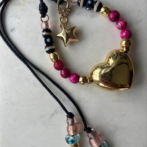 Puede incluir: Un brazalete de cordón negro con cuentas rosas y negras, un dije de corazón dorado, un dije de estrella dorada y un dije de abeja dorada. El brazalete también tiene cuentas azules de ojo turco.