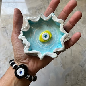 May include: A blue and white ceramic dish with a yellow and blue evil eye design. The dish is in the shape of a flower with a scalloped edge.