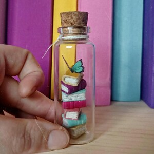Puede incluir: Una pequeña botella de vidrio transparente con un tapón de corcho, que contiene una pila de libros en miniatura de varios colores. Una pequeña mariposa verde descansa sobre los libros. El fondo presenta una fila de libros coloridos.
