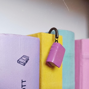 Puede incluir: Un dije de libro miniatura de color púrpura unido a un gancho de metal color bronce, colocado entre un libro lavanda con un icono de libro y el texto COTT, un libro amarillo, un libro azul claro y un libro rosa alineados.
