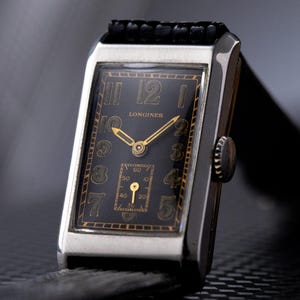 May include: A rectangular wristwatch with a black face and gold numerals. The watch has a silver-colored case and a black leather band. The brand name "LONGINES" is visible on the watch face. The watch is a vintage style.