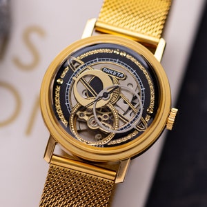 Könnte beinhalten: Goldfarbene Armbanduhr mit einem Mesh-Armband. Das Zifferblatt hat ein schwarzes und goldenes Design mit sichtbaren Innenmechanismen. Das Wort "PAKETA" ist auf dem Zifferblatt aufgedruckt. Die Uhr befindet sich vor einem neutralen Hintergrund.