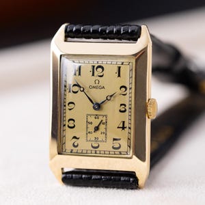 May include: A rectangular gold-toned Omega wristwatch with a cream-colored face and black leather band. The watch face features black numerals and hands, with a small seconds dial. The watch is a classic design.